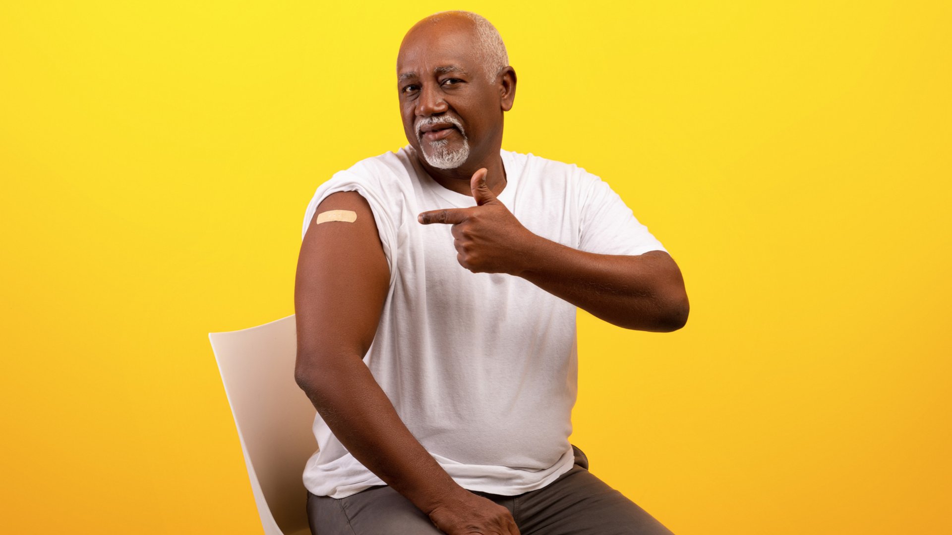 Homem sentado apontando com o dedo indicador esquerdo seu braço direito no qual tem um curativo de vacina