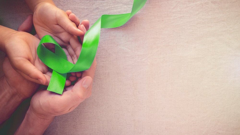Mãos segurando um laço verde claro. Este representa o Agosto Verde Claro, campanha cuja intenção é o combate ao linfoma (câncer da porção linfática) e a conscientização acerca do mesmo.