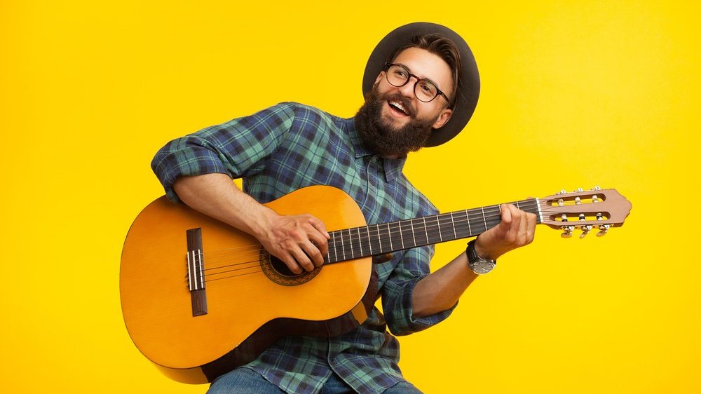 Homem de barba, chapéu e óculos tocando violão em fundo amarelo