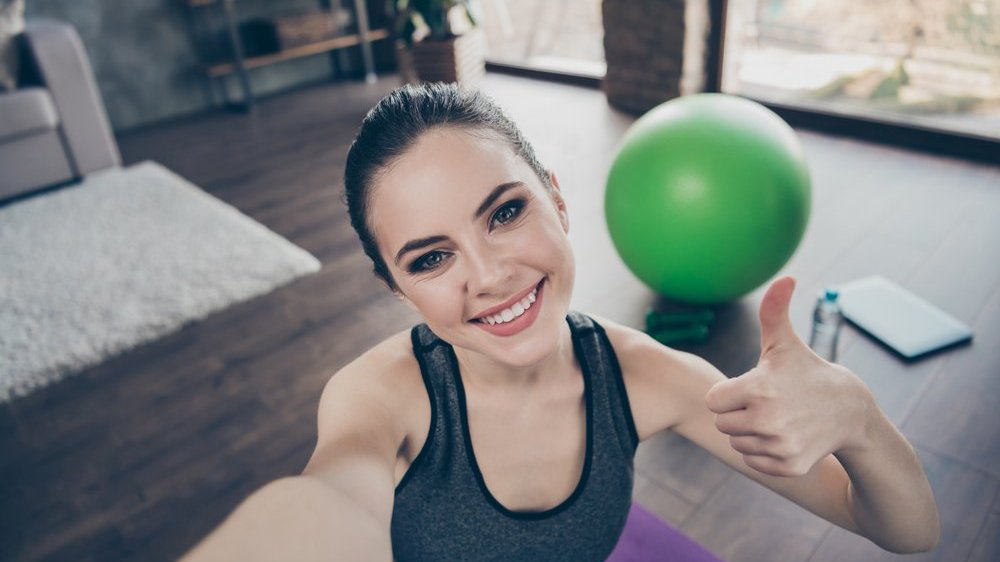 Mulher tirando selfie fazendo um sinal positivo com uma mão e sorrindo enquanto pratica yoga