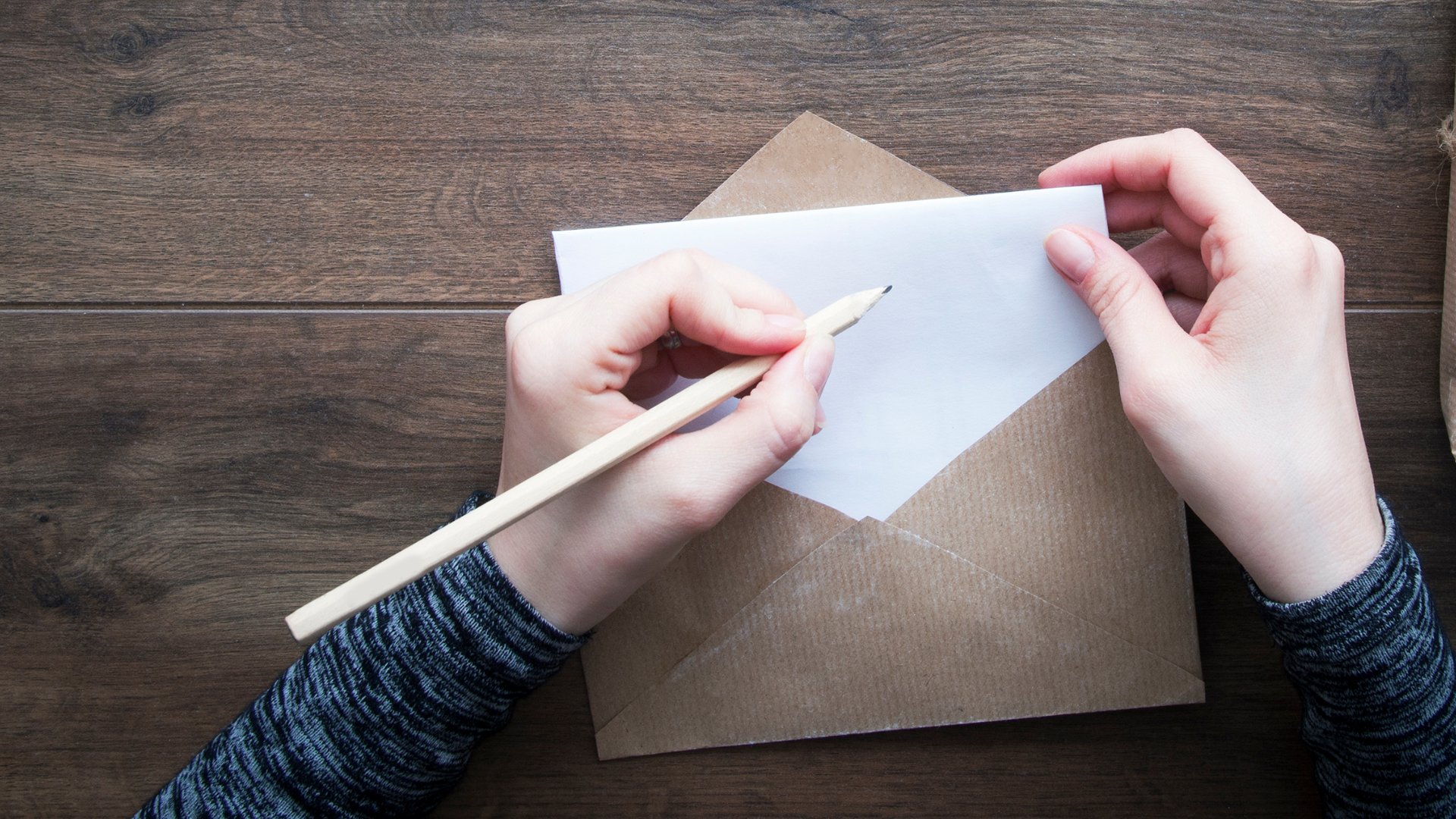Uma pessoa segurando um envelope de carta. Na carta, propriamente, a mão esquerda dessa pessoa escreve algo com um lápis.