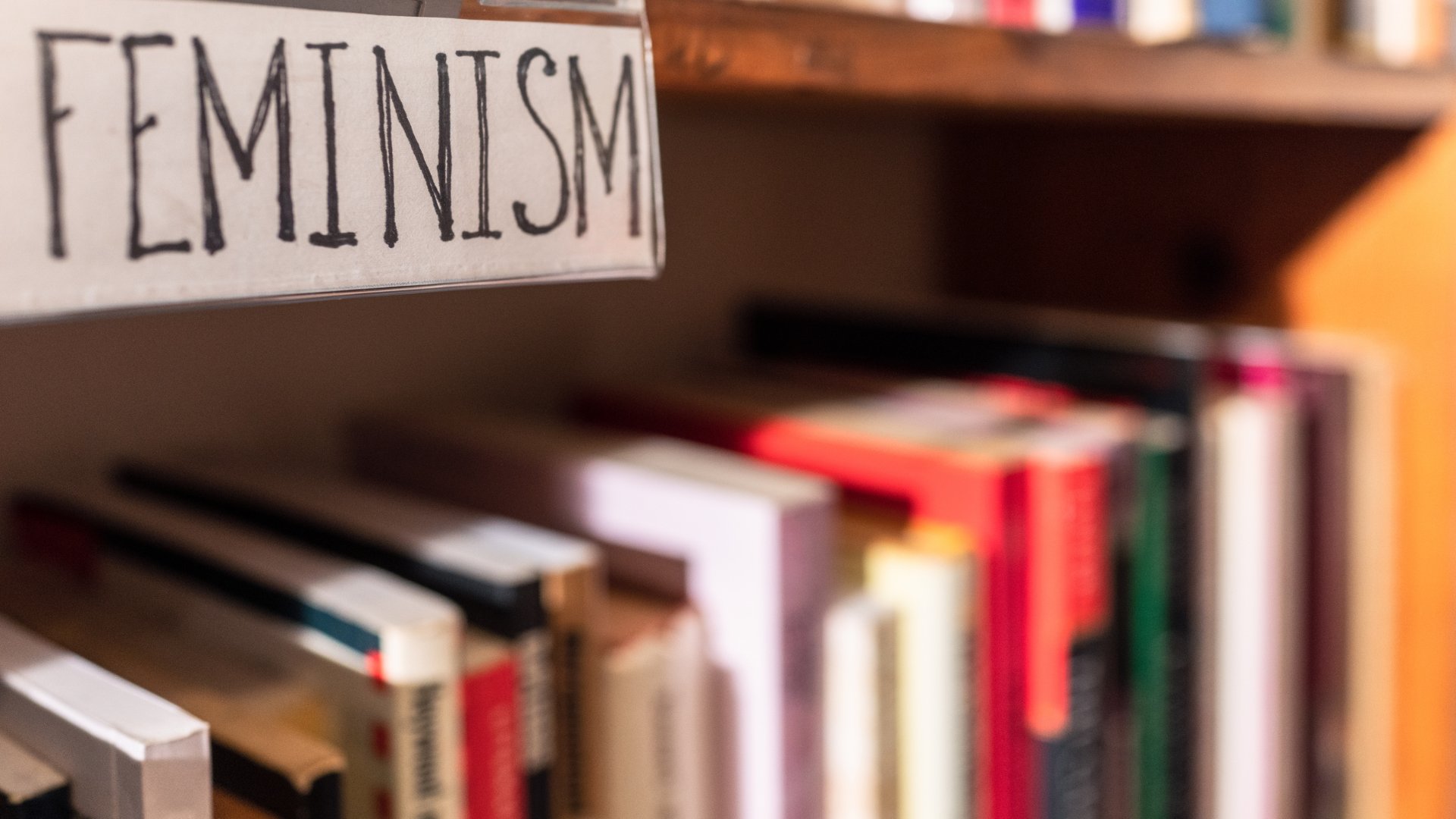 Uma prateleira de livros e, acima desta, uma placa cujo escrito presente é a palavra 'feminism'