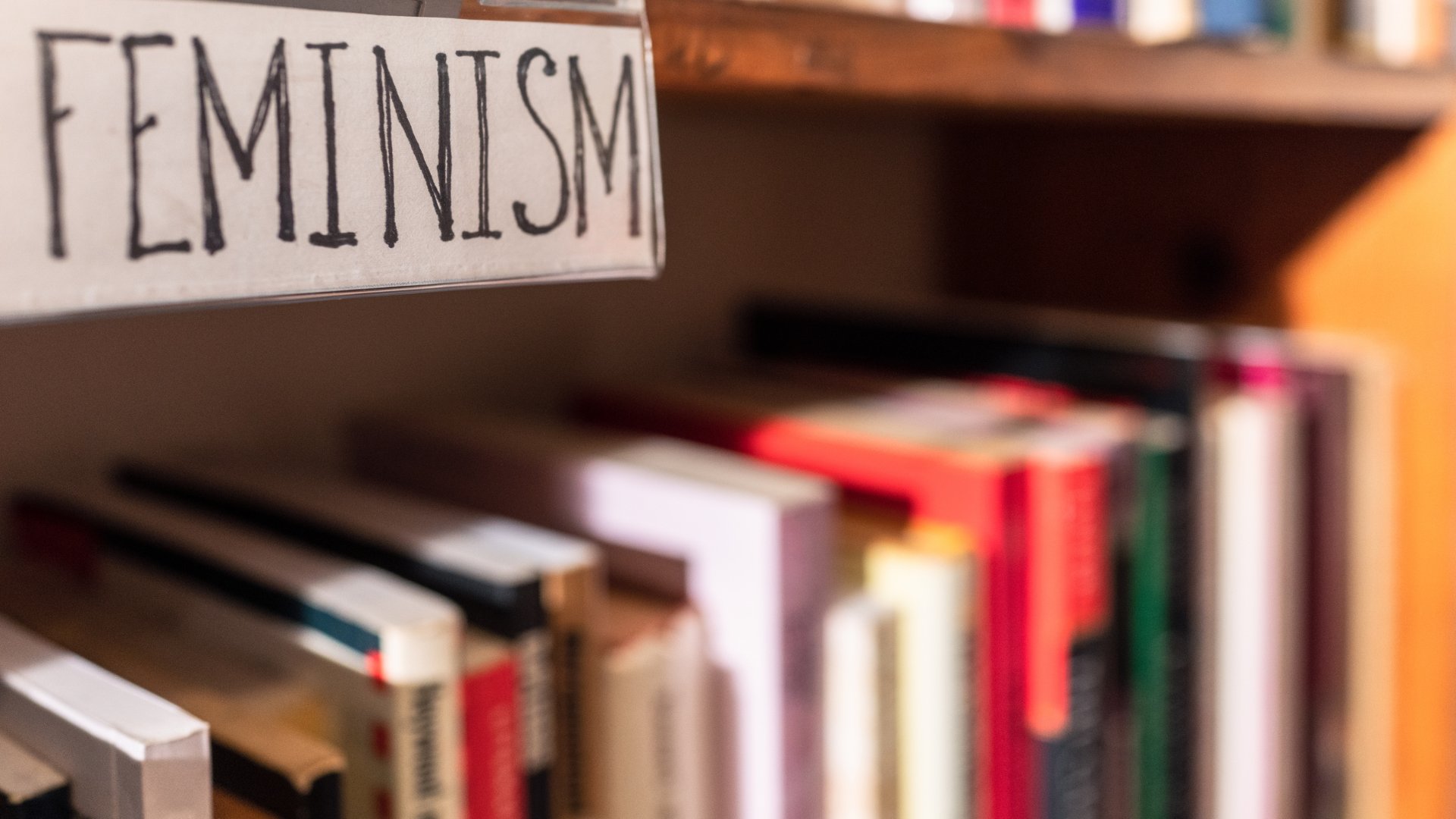 georgeclerk de Getty Images Signature / Canva Uma prateleira de livros e, acima desta, uma placa cujo escrito presente é a palavra 'feminism'
