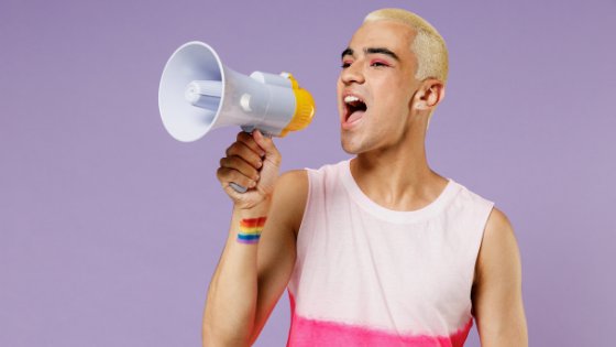 Adolescente segurando e falando ao megafone, com a bandeira LGBTQIA+ desenhada em seu pulso.