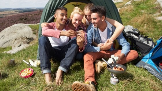 Duas mulheres e dois homens jovens, sentados em frente a uma barraca de acampamento, sorrindo ao olhar o celular.
