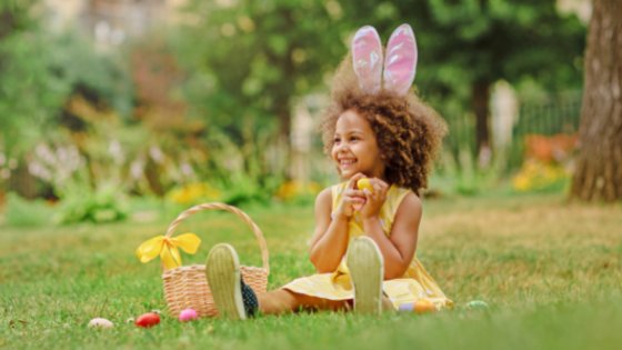 Menina sentada na grama ao lado de uma cesta de ovos de chocolate, com orelhas de coelho