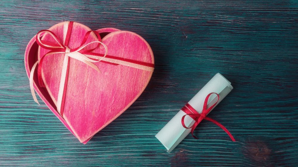 Caixa em formato de coração rosa e bilhete de papel enrolado com laço sobre uma mesa