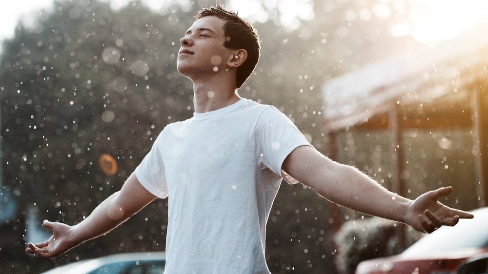 Homem na chuva com os braços abertos e olhos fechados