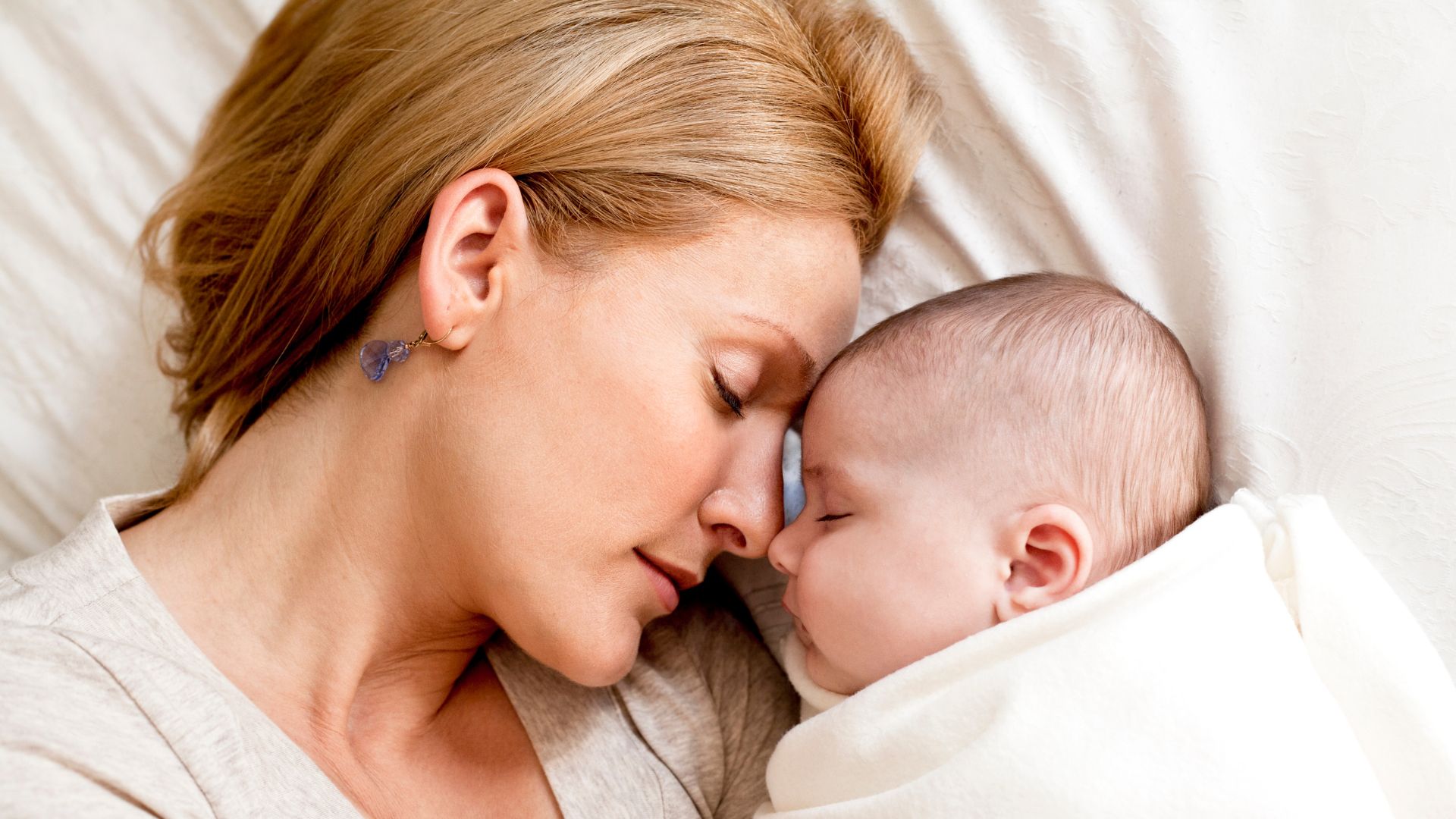 Uma mulher branca de cabelos loiros encostando sua cabeça na cabeça de um pequeno bebê.