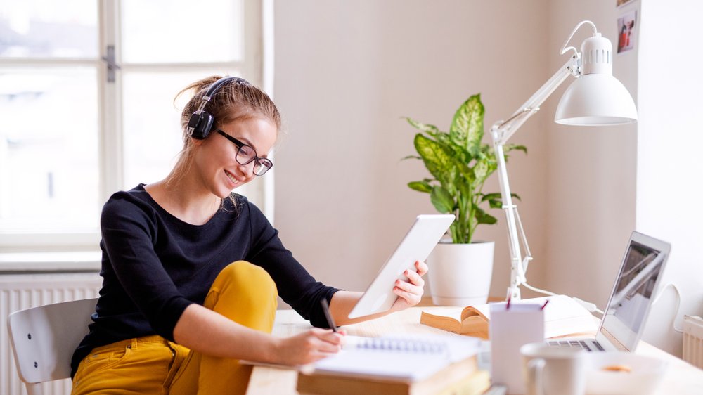 Mulher de fones de ouvido sentada à mesa estudando enquanto está sorrindo