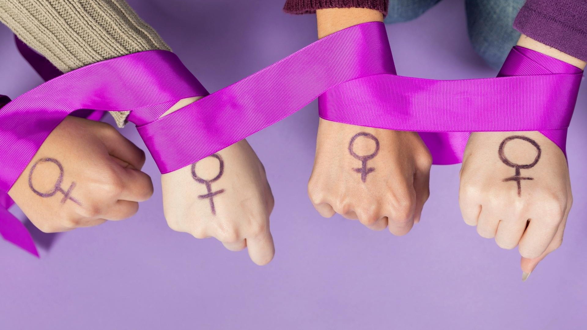 Um fita percorrendo pulsos de várias mulheres. Na fronte da mão de cada uma das mulheres, um símbolo que representa o sexo feminino.