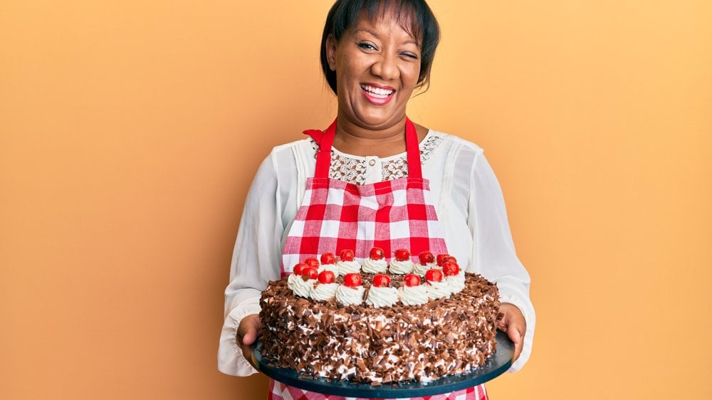 Mulher negra com avental de confeiteira segurando um bolo de aniversário