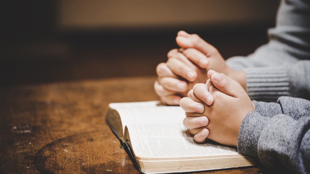 Sobre uma Bíblia aberta, mãos de um adulto e de uma criança cruzadas em forma de oração.