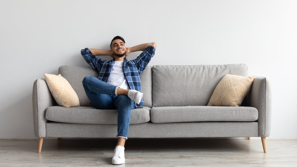 Um homem branco sentado num sofá. Ele ergue seus braços e os apoia atrás da sua cabeça.