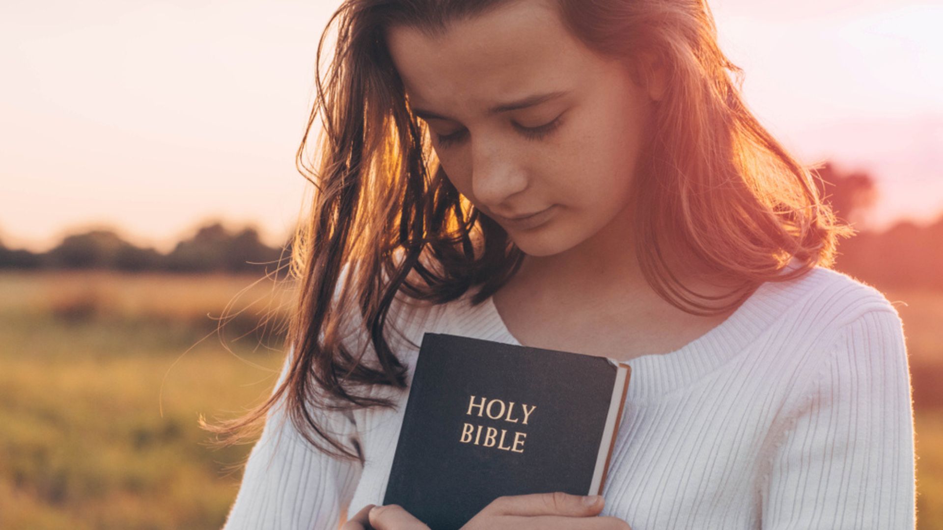 Uma mulher branca de visagem baixa e olhos fechados. Ela segura um exemplar da Bíblia contra o peito.