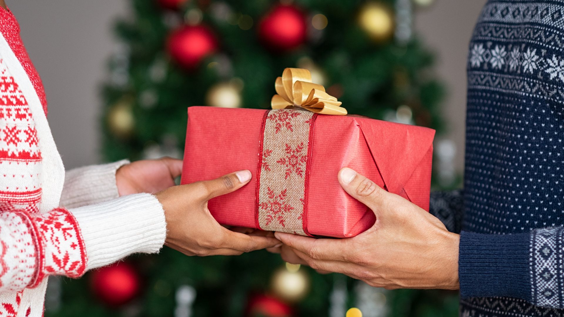 Imagem de uma mulher entregando um presente de natal para um homem e ao fundo uma árvore de Natal