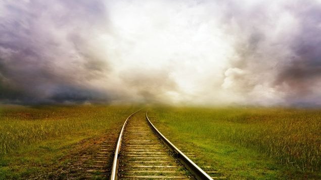 Trilho de trem atravessando por um campo florido e, no fim, por uma paisagem cinzenta