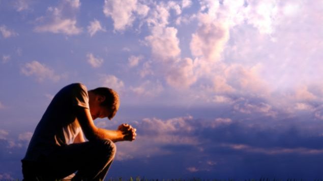 Homem abaixado, rezando, com o céu azul atrás.
