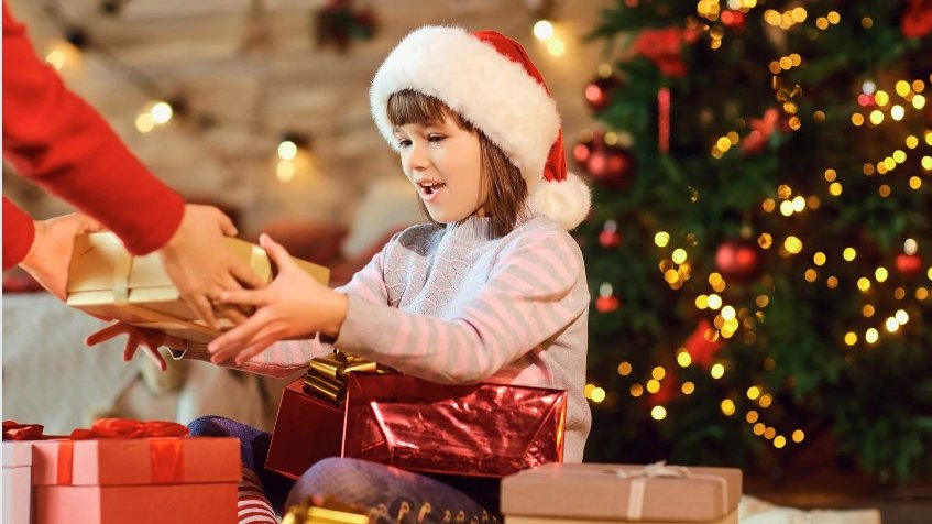 Criança recenedo um presente de natal, vestida com uma touca de natal, com uma árvore decorativa atrás