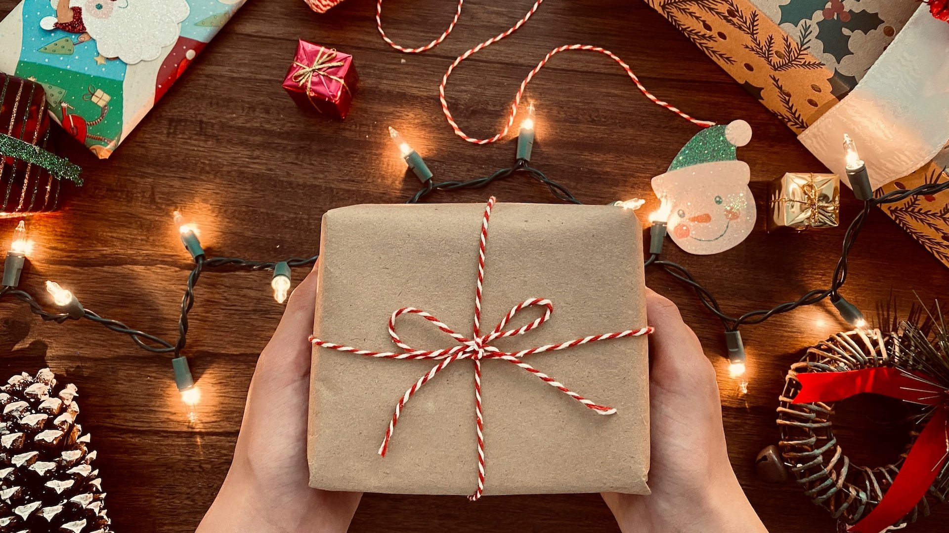 Mãos segurando uma embalagem simples de presente sobre uma mesa cheia de decorações de Natal.
