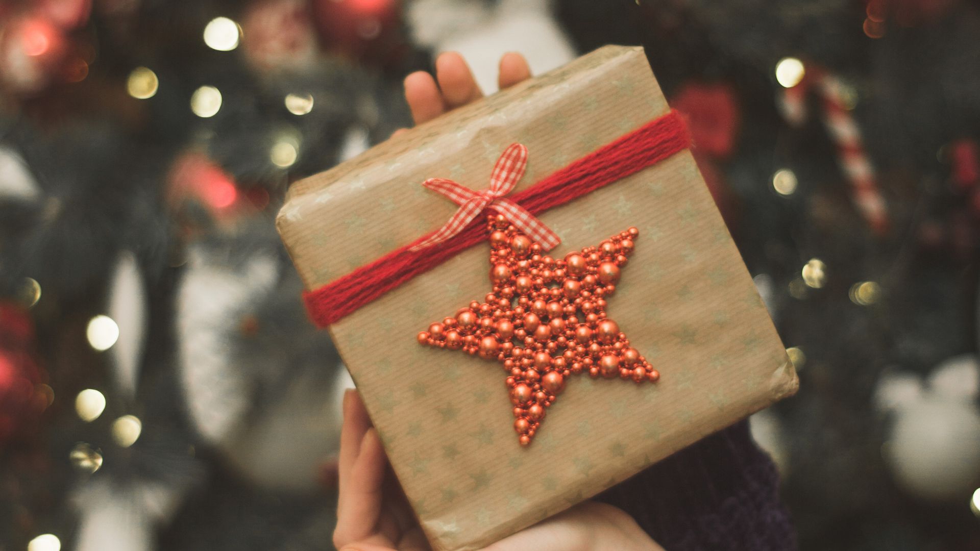 Imagem de um presente de natal em uma embalagem bege e uma estrela vermelha de enfeite na frente, e ao fundo luzes em uma árvore de natal