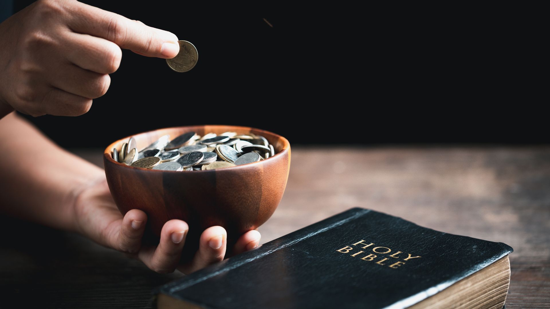 Imagem de uma bíblia em cima de uma mesa e ao lado uma pessoa fazendo oferta de moedas em um potinho de barro
