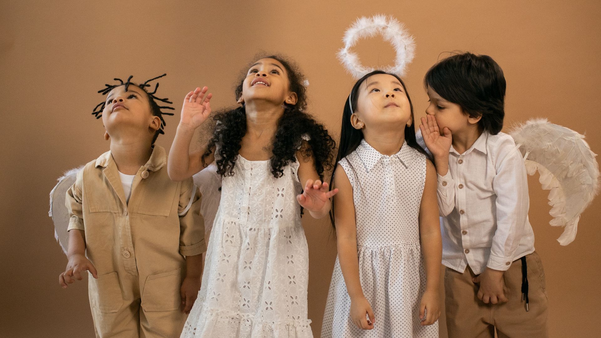 Imagem de quatro crianças em um fundo bege, vestidas de anjinhos, três olhando para cima e uma falando no ouvido da terceira