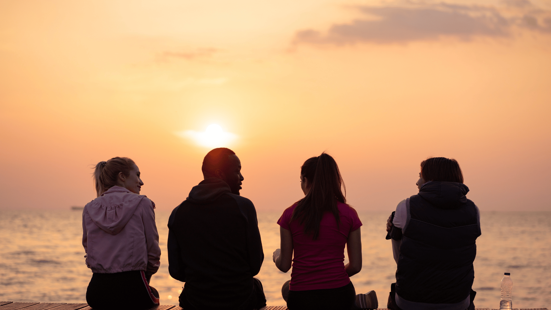 Três mulheres e um homem sentados observando o pôr do sol a beira do mar.