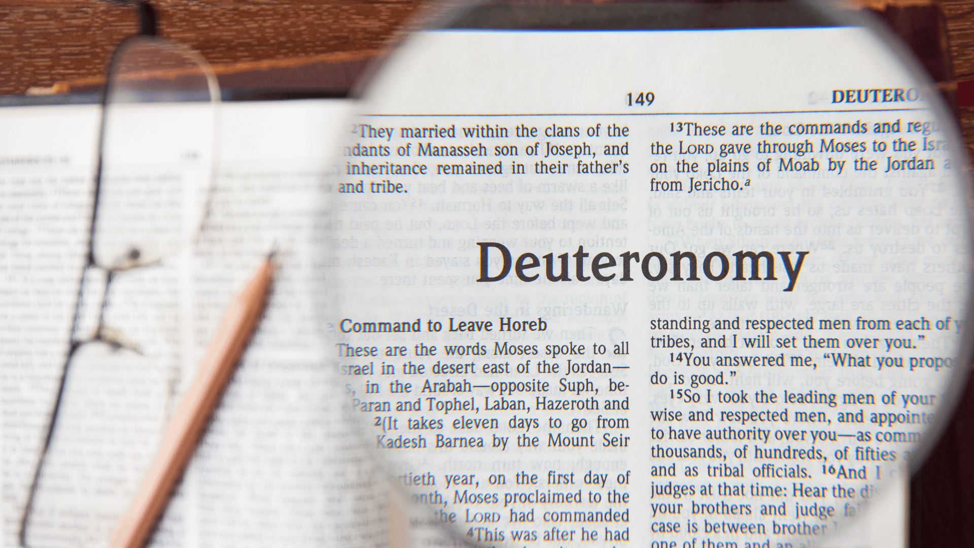 Livro de Deuteronômio em língua inglesa posicionado em uma mesa com um óculos e um lápis ao lado. O nome 'Deuteronômio' está focado por meio de uma lupa.