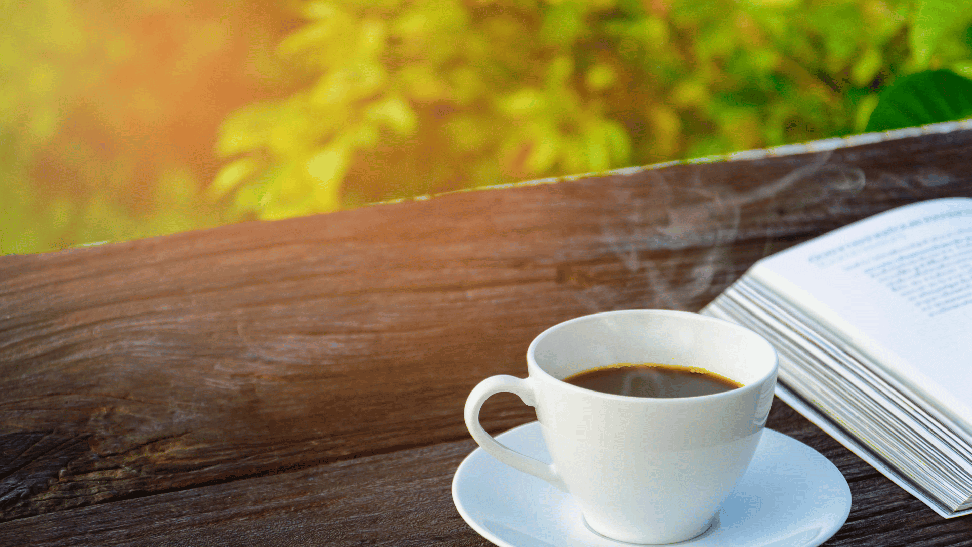 Uma xícara branca com café ao lado de um livro, ambos em uma mesa de madeira em um jardim com plantas.