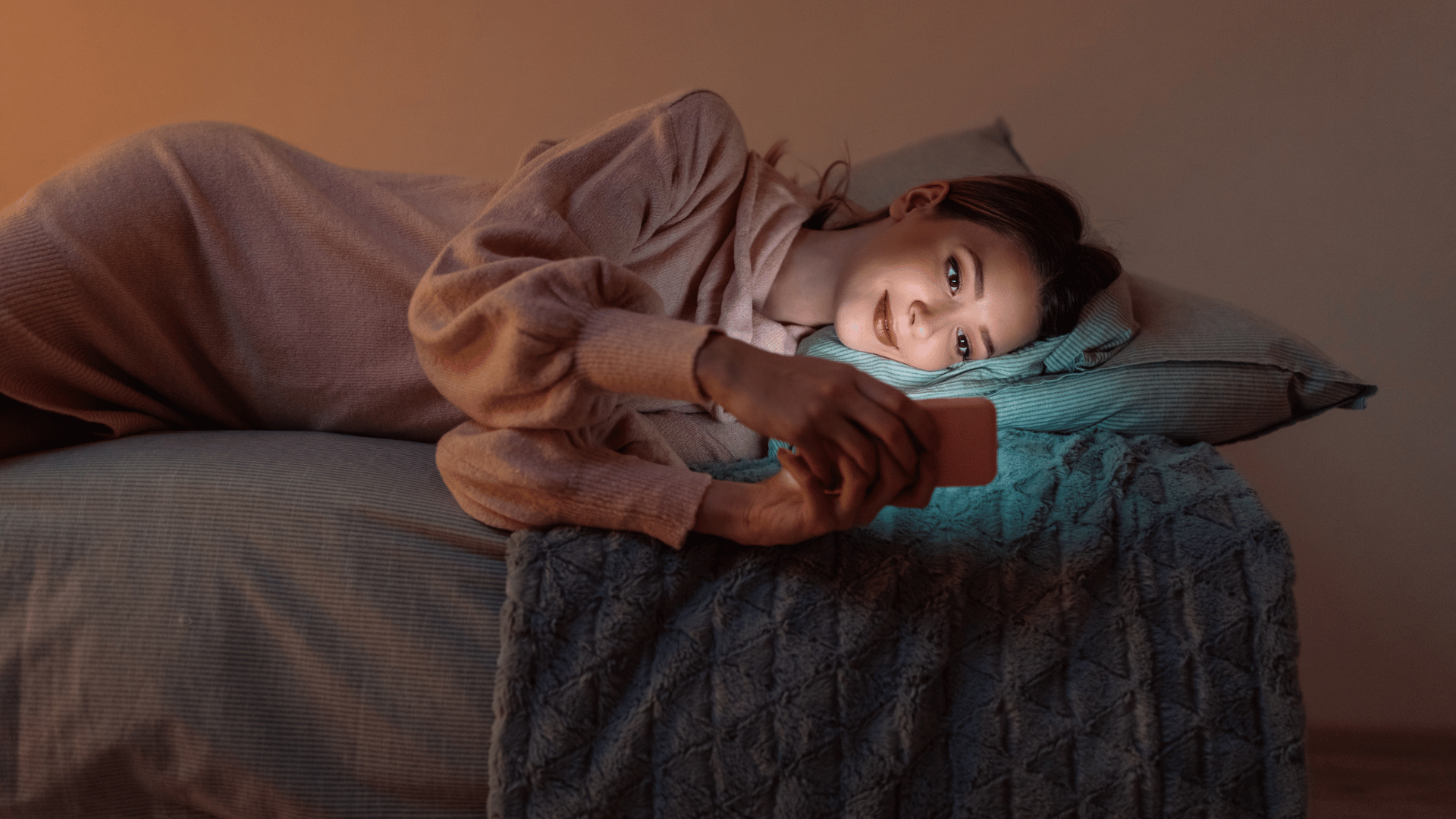 Mulher branca deitada na cama sorrindo enquanto mexe no celular.