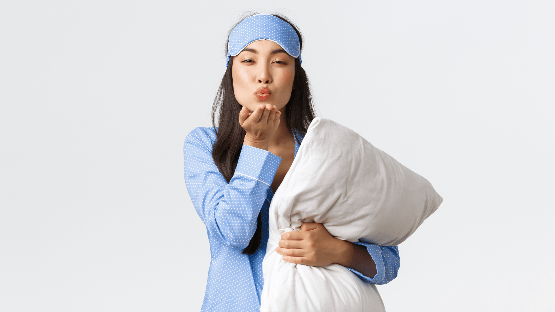 Garota asiática vestida com um pijama azul e máscara de dormir, segurando um travesseiro enquanto faz sinal de beijo.