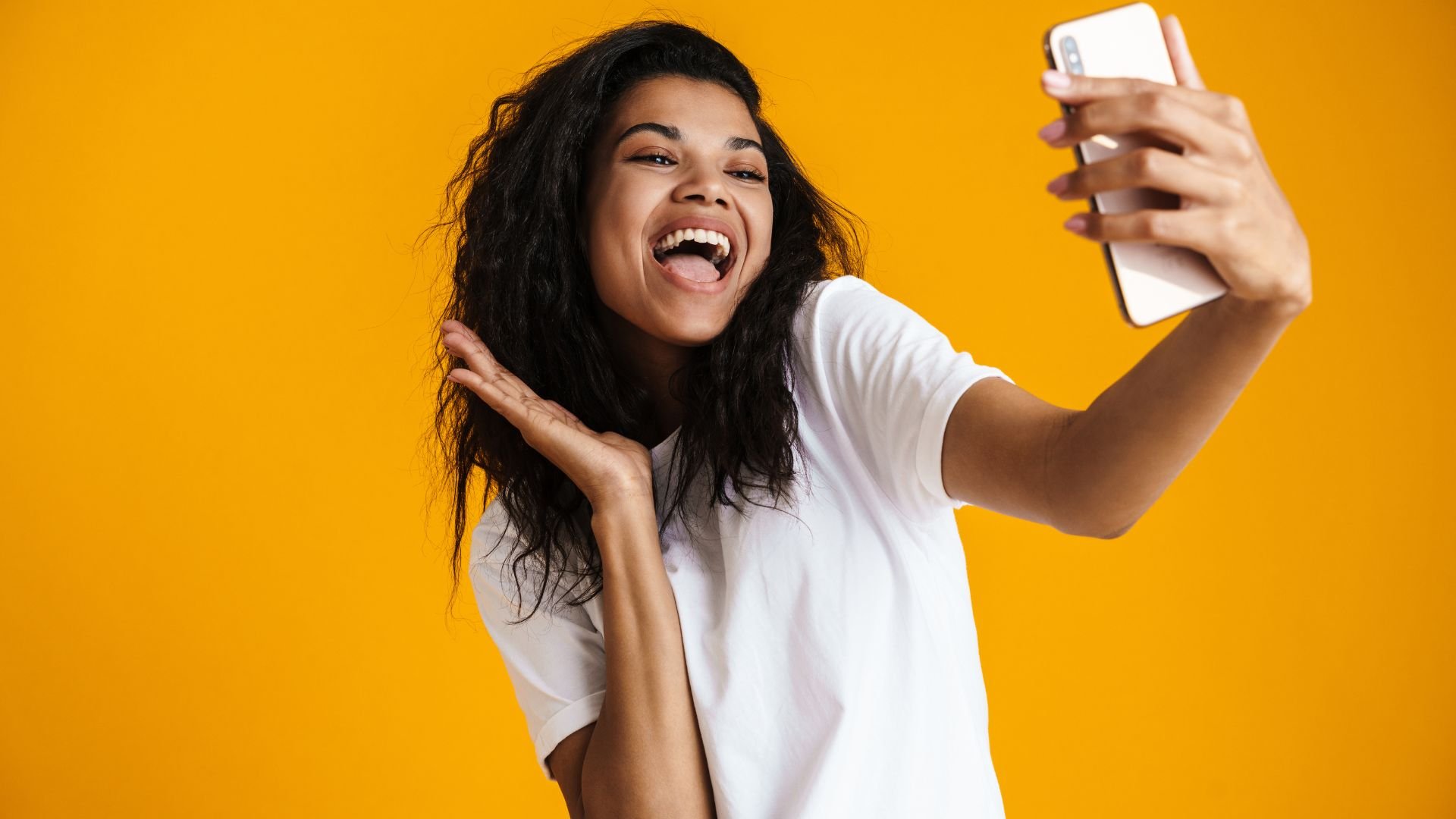Imagem de uma mulher sorrindo e capturando uma foto selfie com o celular em um fundo amarelo