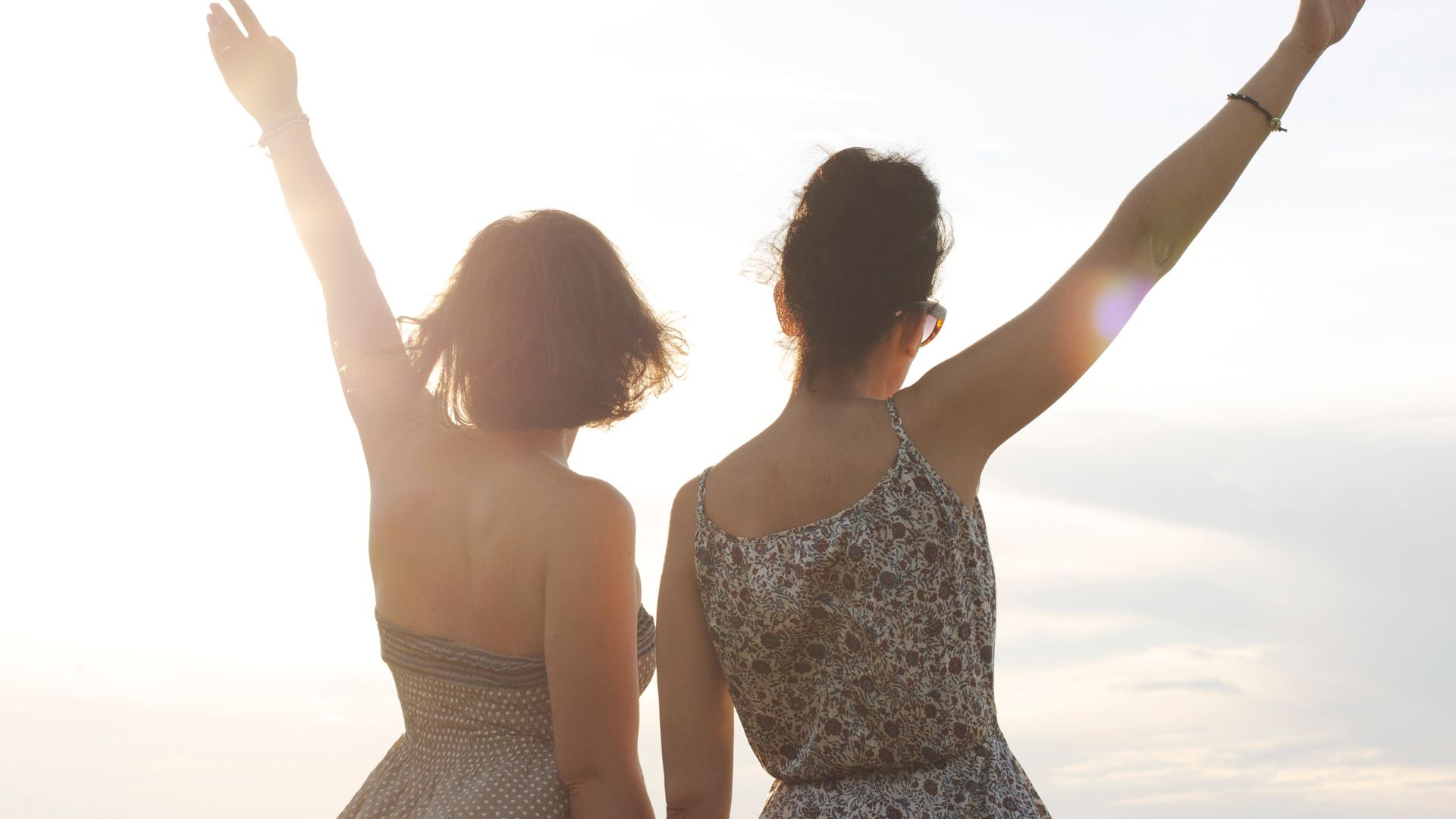 Imagem de duas amigas no topo de uma montanha com os braços levantados contemplando o sol