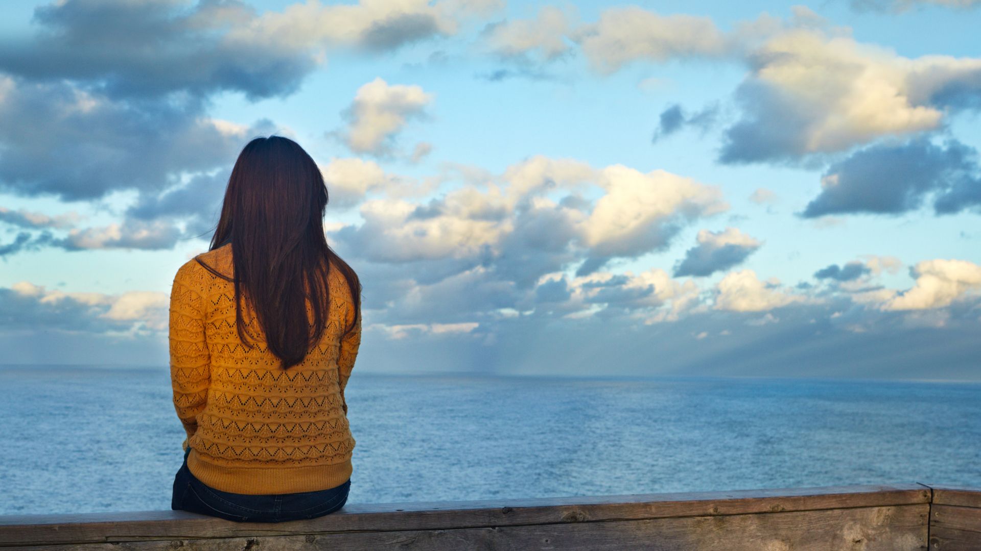 Menina sozinha e sentada diante um céu azul com nuvens.