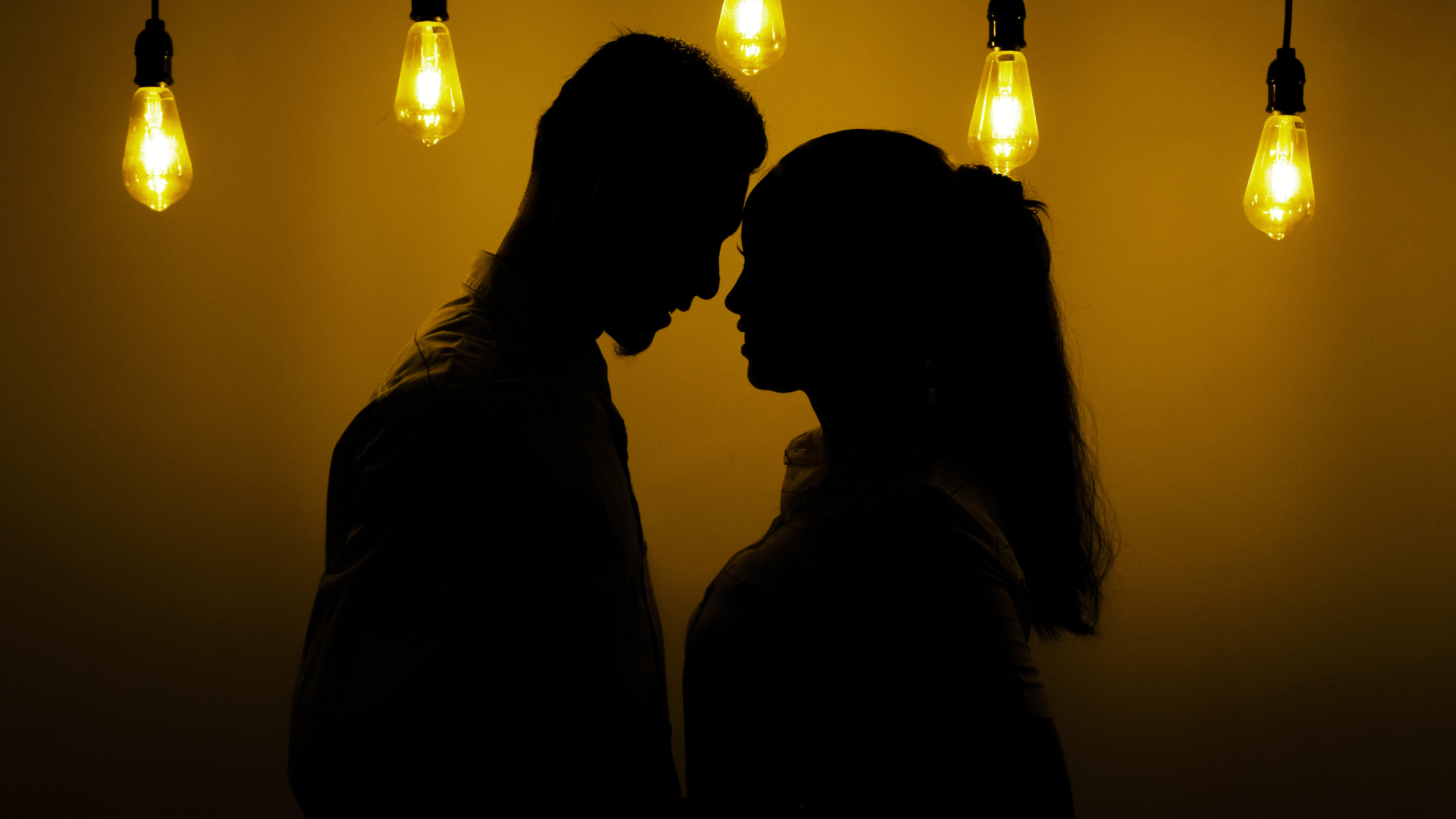 Silhueta de um casal romântico com as faces próximas, em um cenário amarelo escuro com luzes.