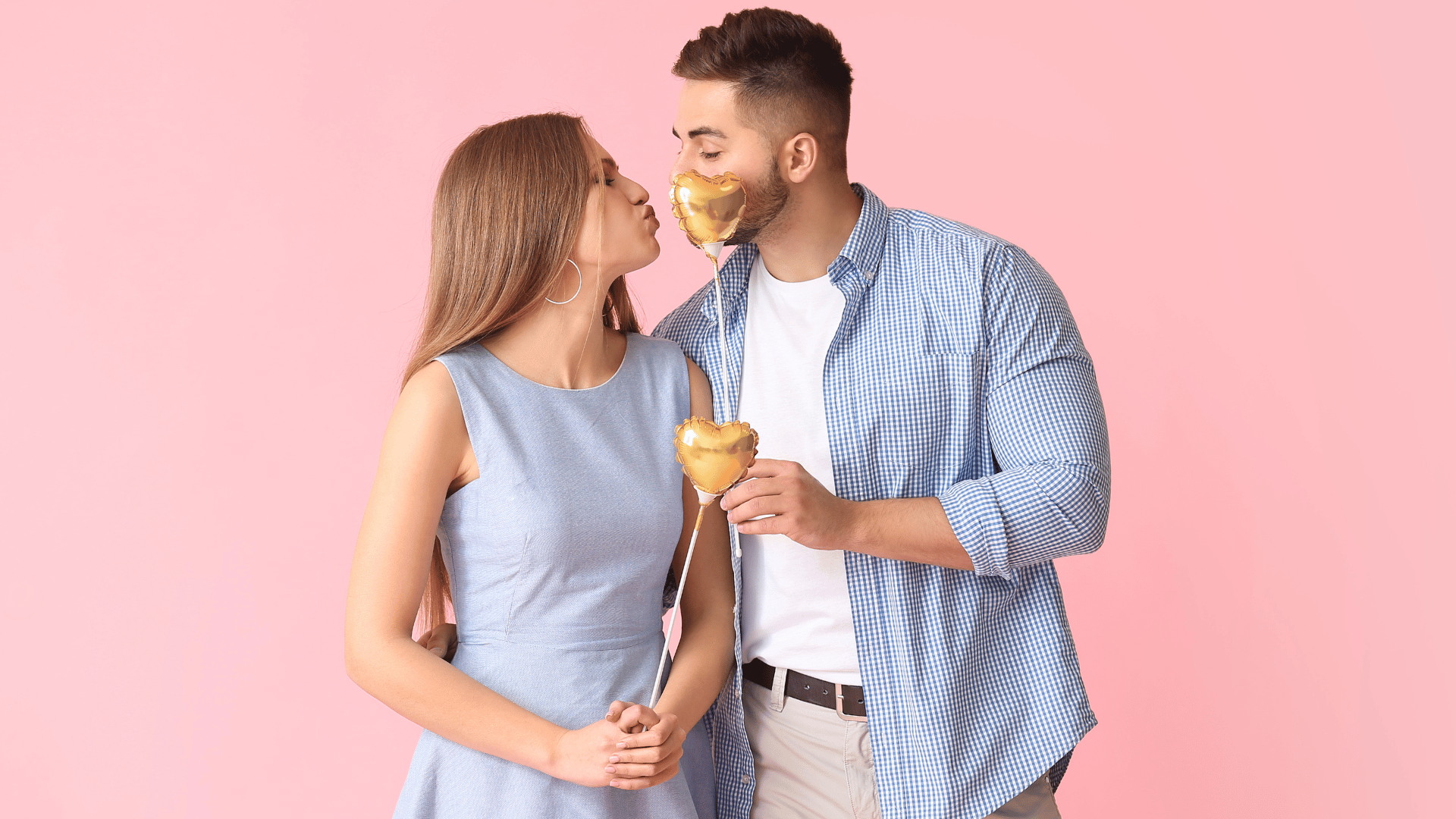 Casal jovens segurando bexigas pequenas em formato de coração, em um fundo rosa bebê.