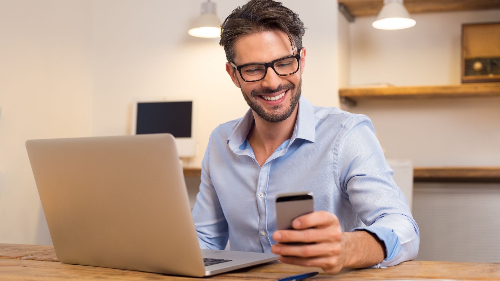 Homem com um laptop em sua frente e rindo com o celular na mão