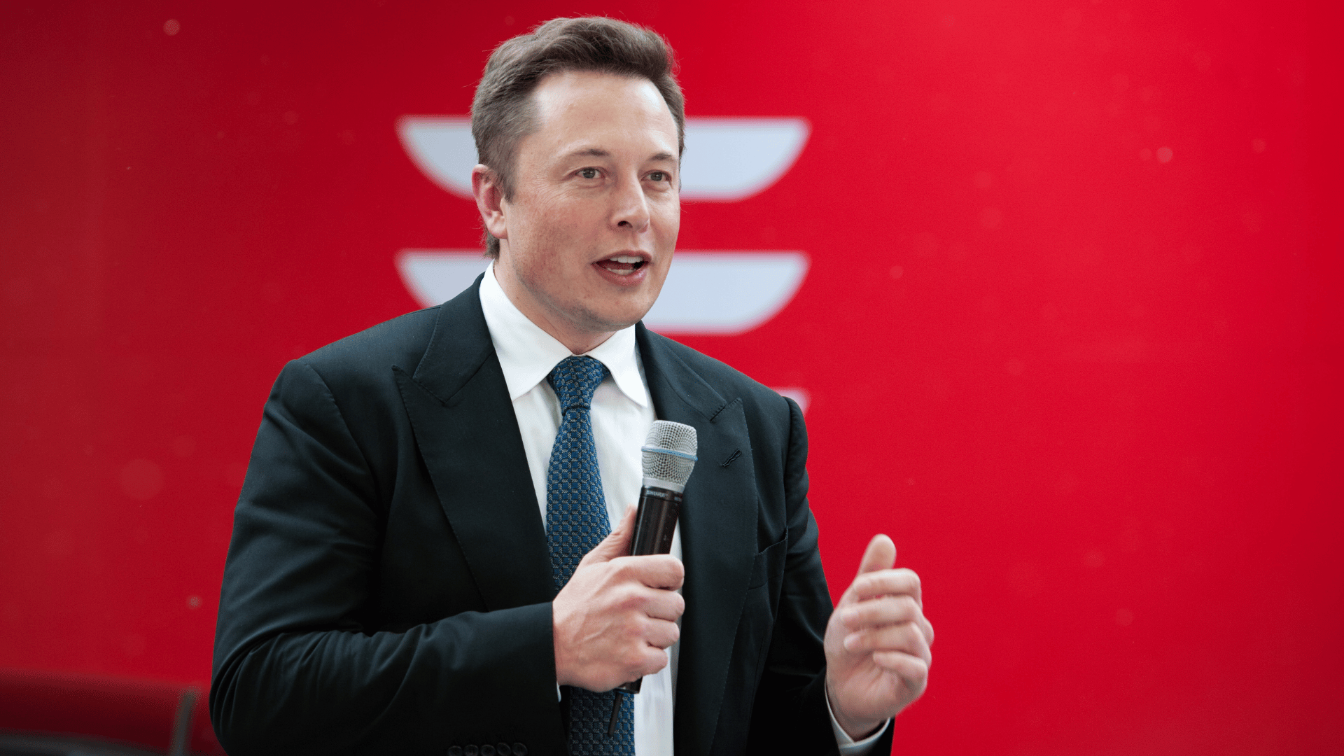 Imagem de Elon Musk. Ele segura um microfone e está sobre um fundo vermelho.