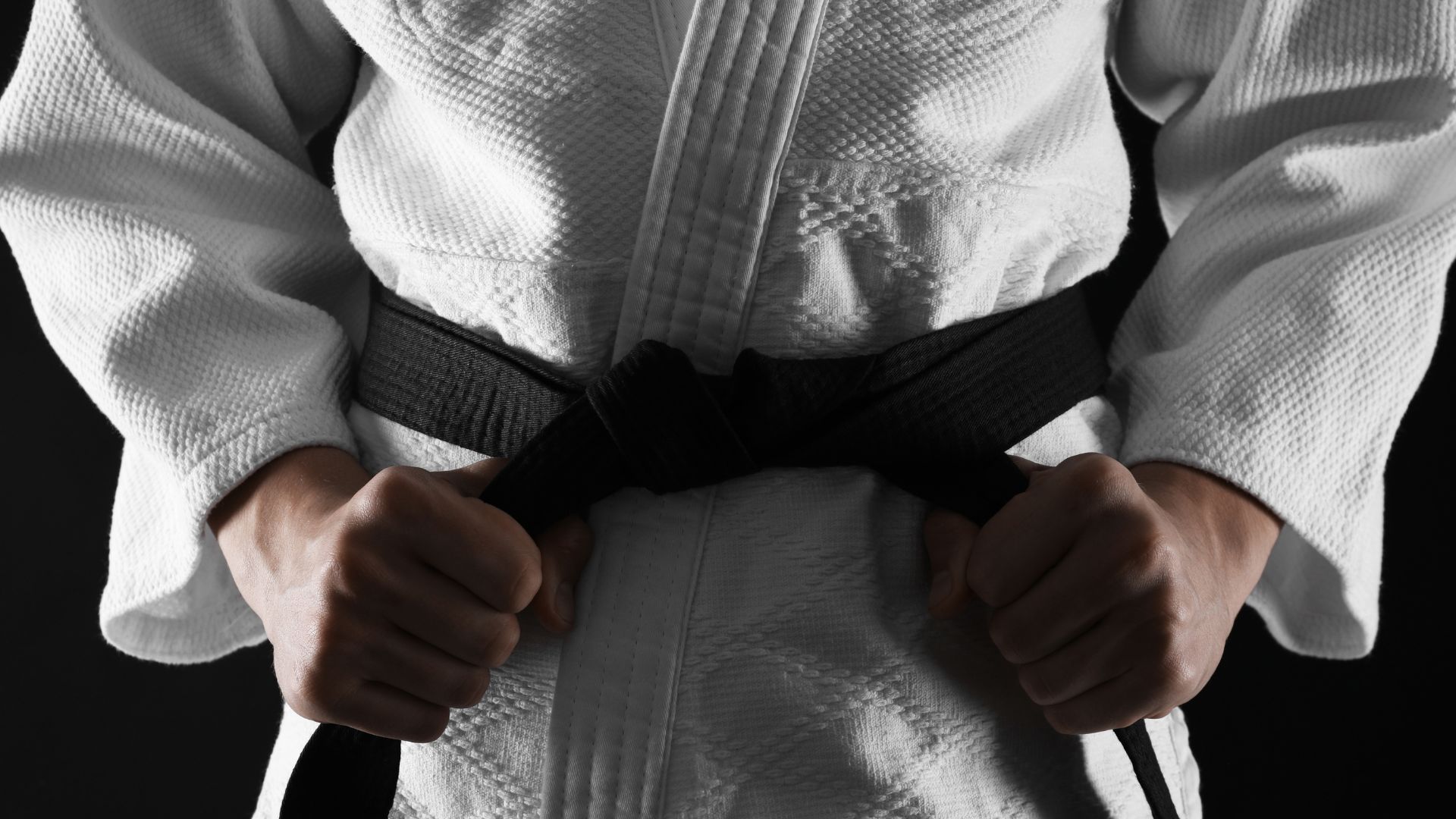 Homem em Keikogi amarrando faixa preta em fundo escuro. Conceito de jiu-jitsu.