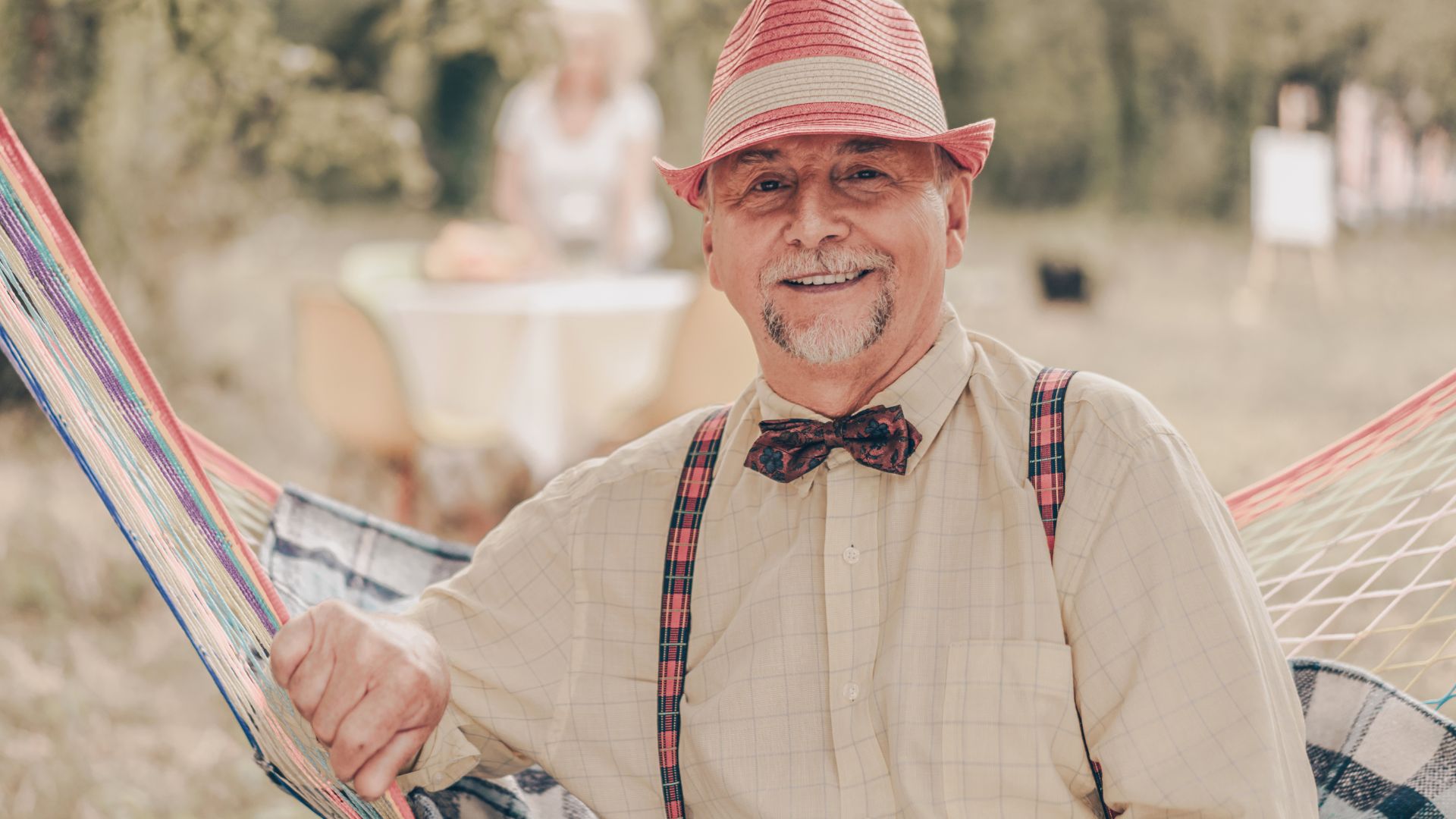 Imagem de um idoso, sentando em uma rede de balanço. Ele está sorrindo, usa um chapéu rosa, uma camisa bege xadrez, suspensórios e gravata borboleta.