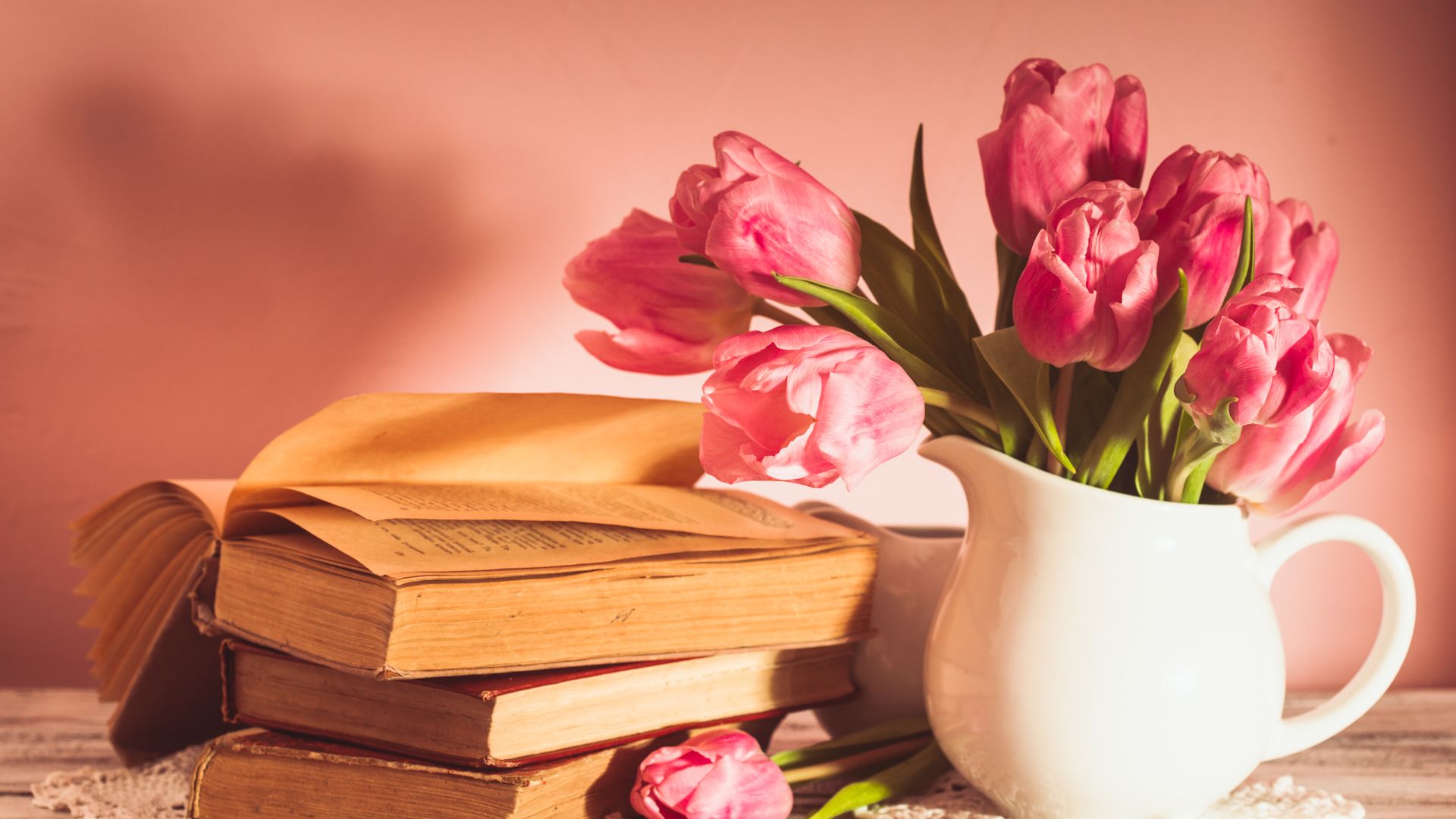 Imagem de fundo rosa e em destaque um livro aberto sobre poemas da vida, um vaso branco com várias tulipas na cor rosa claro.