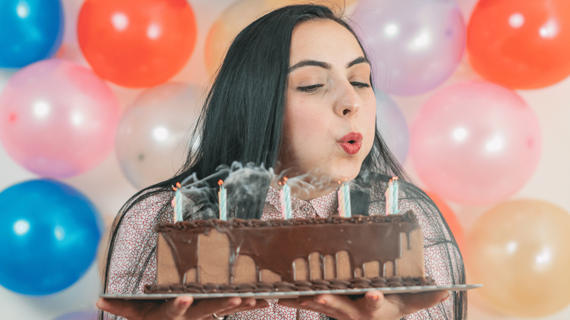 Uma mulher jovem está segurando seu bolo de aniversário. Ela apaga as velas que estão em cima do bolo. No fundo, há balões.